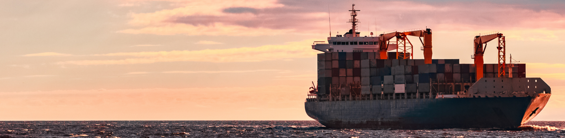 Large cargo ship with containers on board sailing on the ocean at sunset, highlighted with orange and pink hues in the sky and reflections on water.