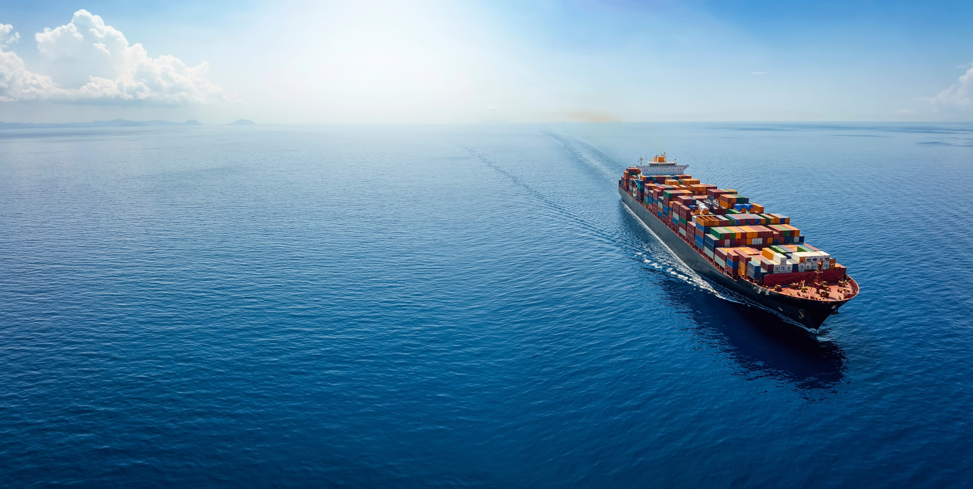 A large cargo ship loaded with colorful containers sailing across a calm blue ocean under a clear sky, with sunlight reflecting on the water.