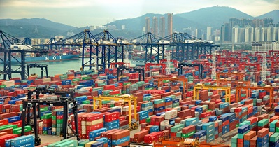 Busy container port with numerous colorful shipping containers stacked up, cranes, and ships, with a backdrop of a city skyline and mountains.