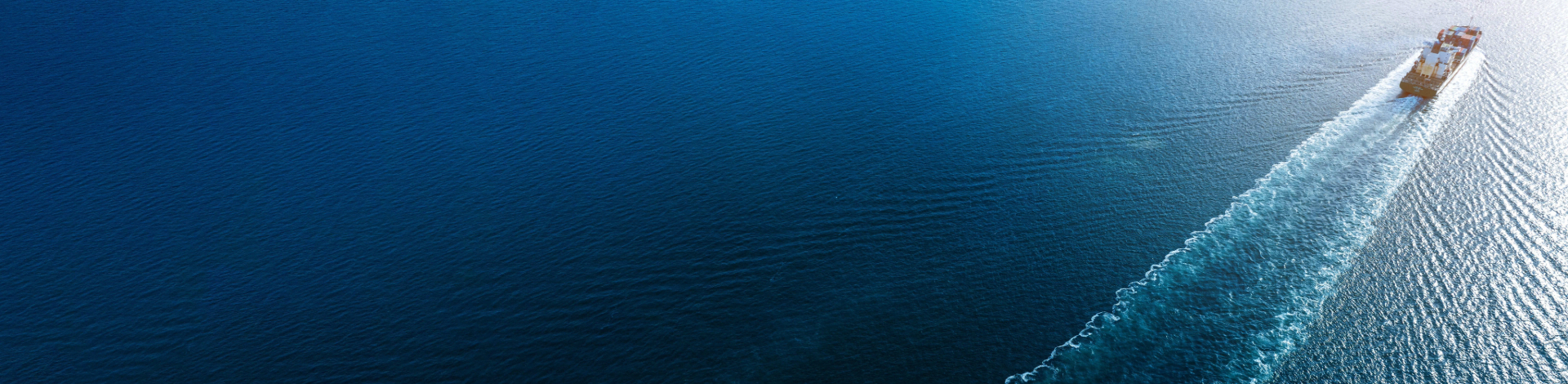 Aerial view of a large cargo ship traveling through a deep blue sea, leaving a long, white wake behind it.