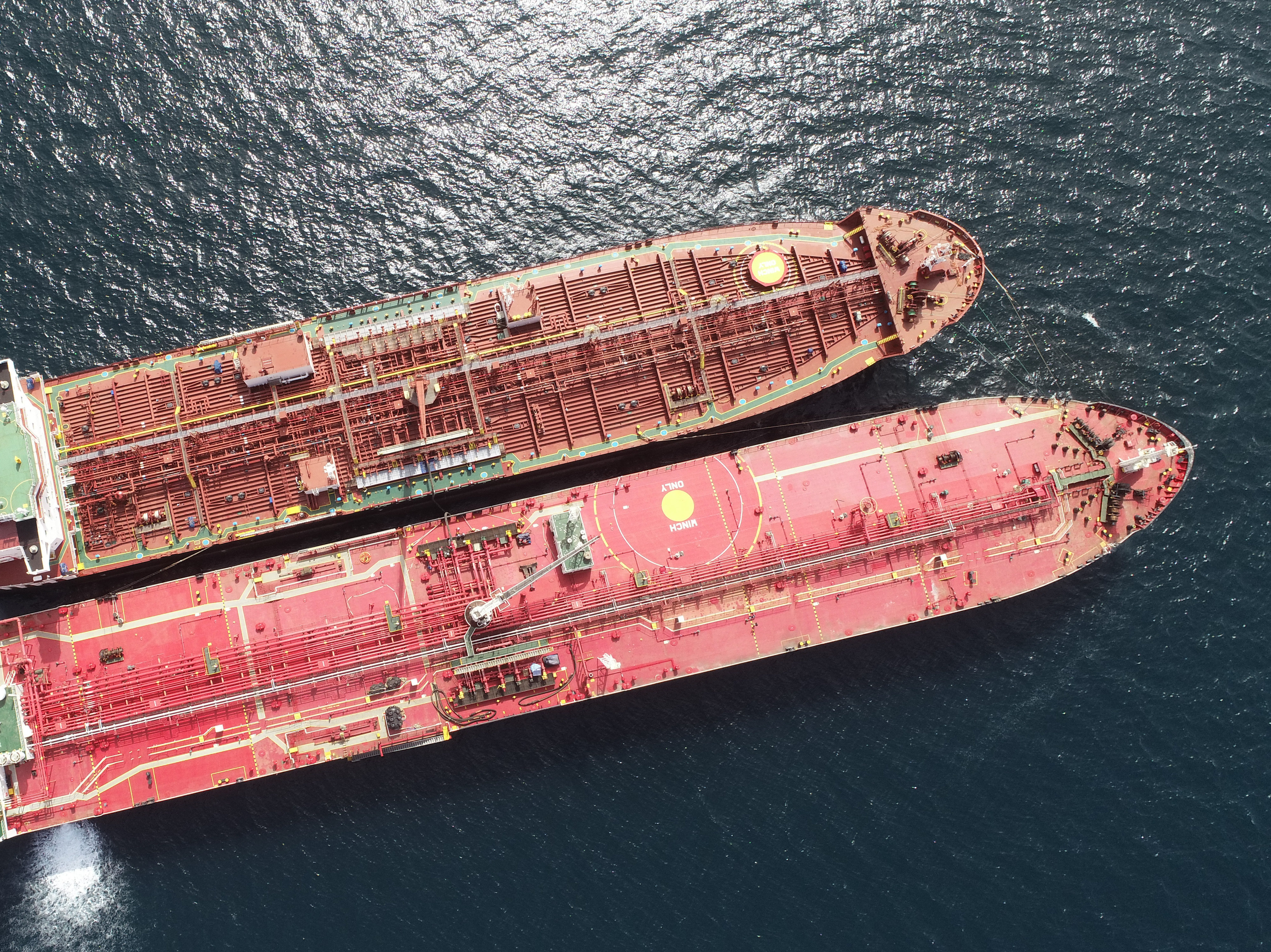 An aerial view of two large oil tankers positioned side by side on a calm blue ocean, showcasing their intricate deck pipelines and operational structures.
