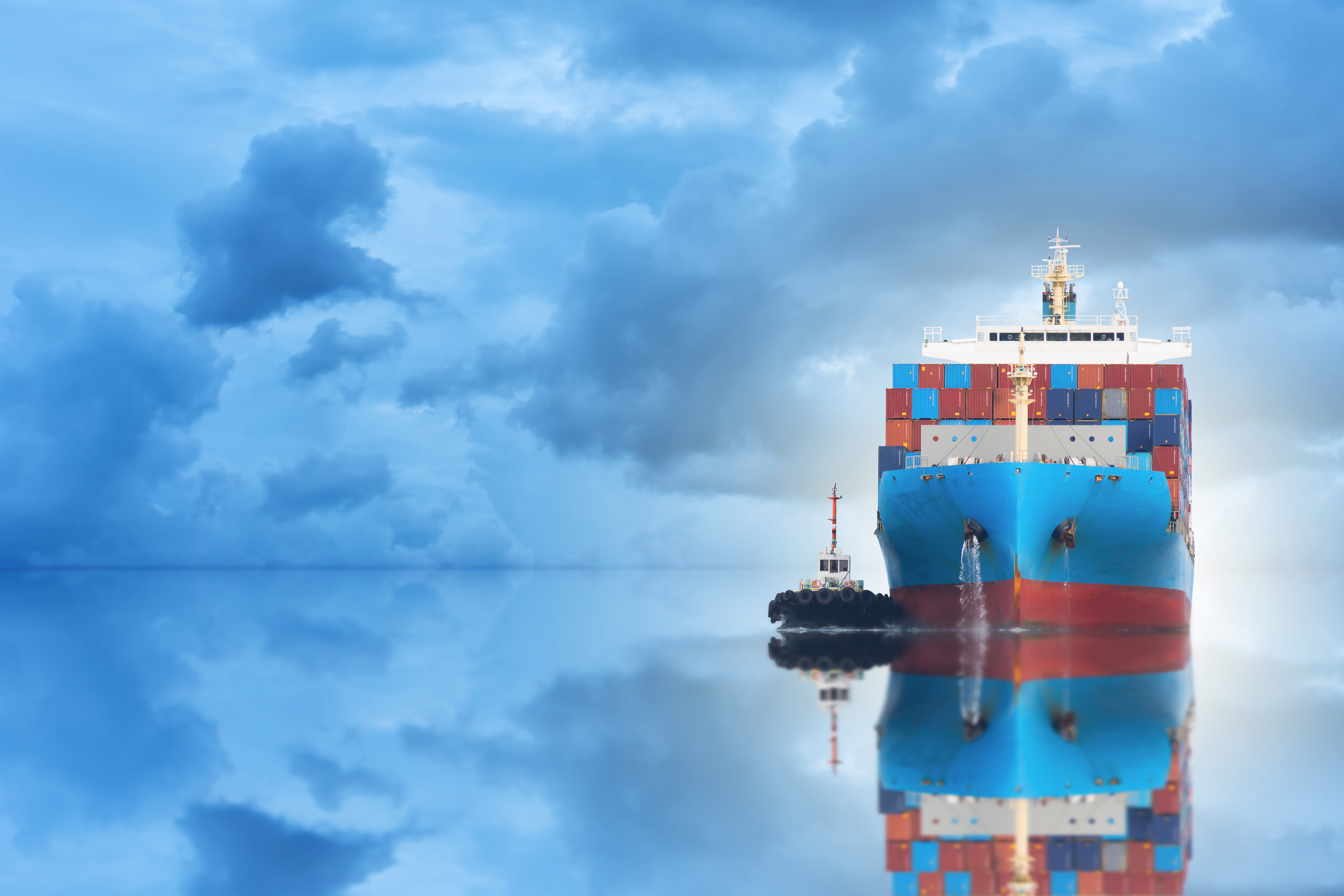 A vibrant front-facing view of a large blue cargo ship stacked with colorful shipping containers, accompanied by a small tugboat on calm reflective waters under a dramatic cloudy sky