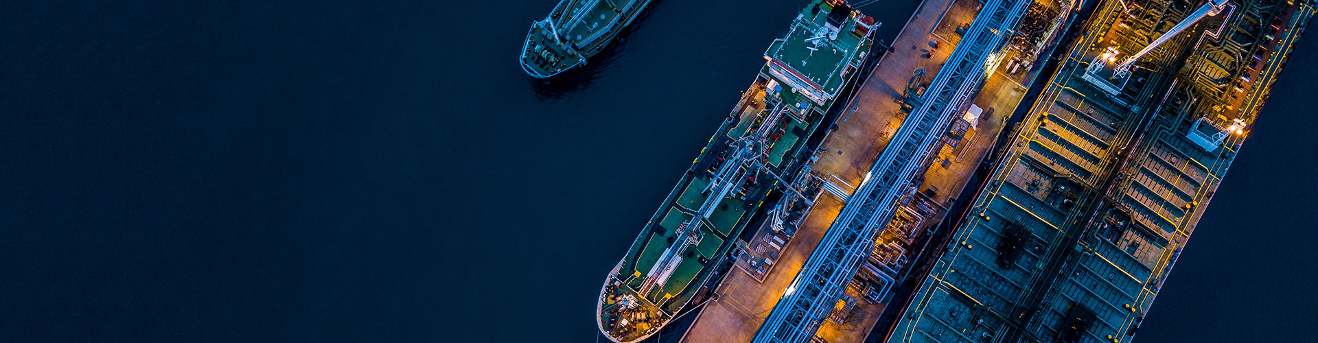 Aerial view of a brightly lit industrial port at night with two large ships docked and numerous containers and equipment visible.