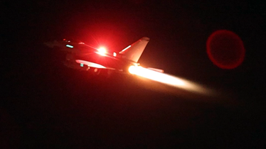 A fighter jet soaring through the night sky, illuminated by its bright lights.