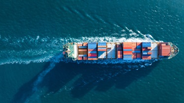 Aerial view of a large cargo ship carrying stacked shipping containers on the ocean, leaving a wake behind it.