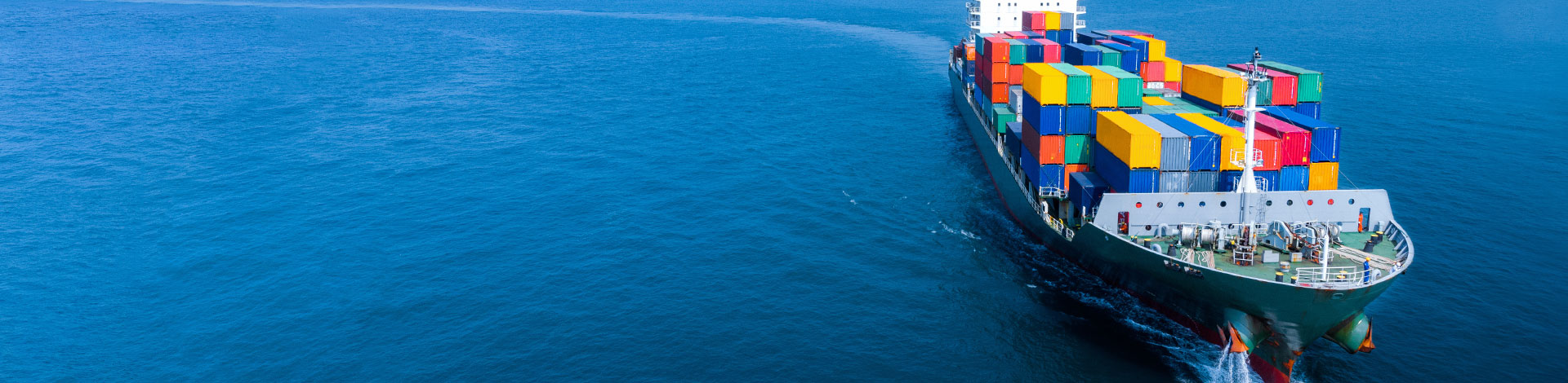 Aerial view of a large cargo ship in the ocean, carrying numerous colorful shipping containers, moving through blue water, creating a wake.